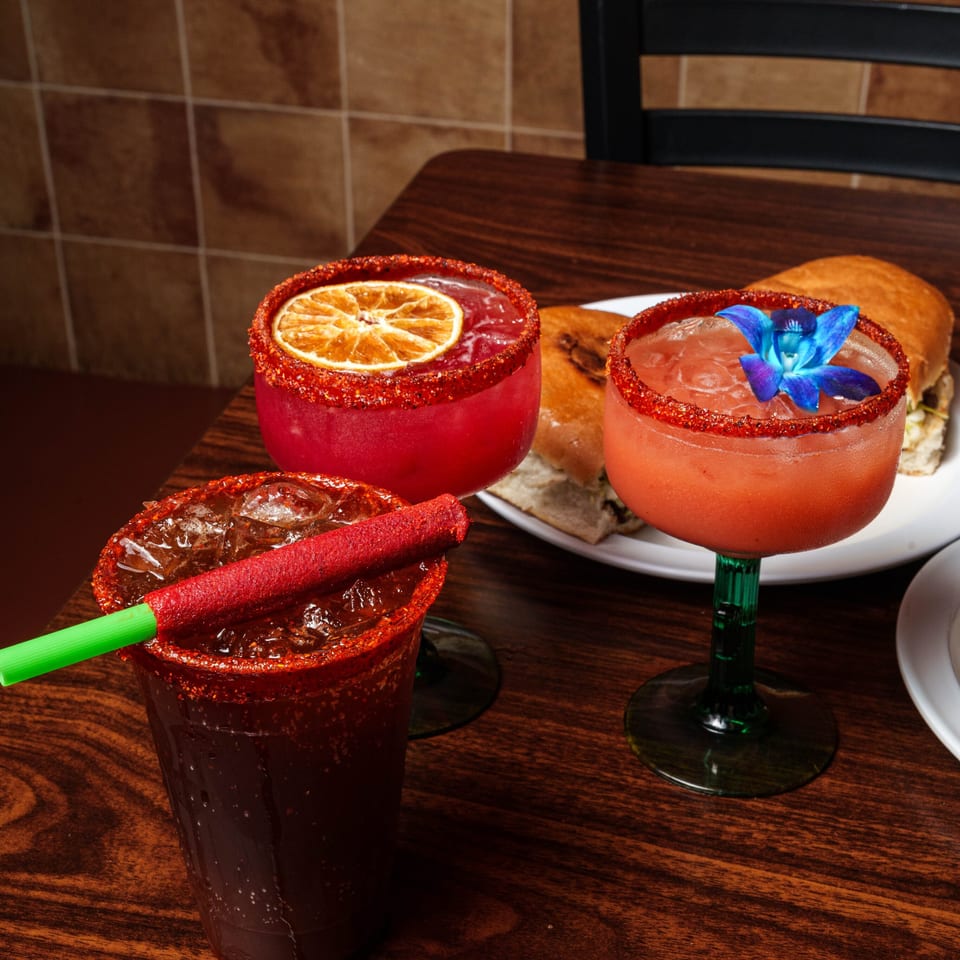 Three vibrant chili-rimmed drinks, including margaritas, and sandwiches on a dark wooden table.