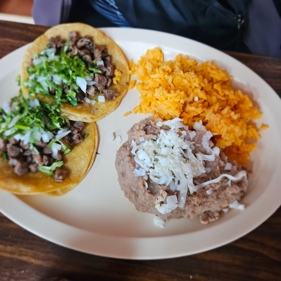 Two street tacos with meat, cilantro, and onions, served with orange rice and refried beans topped with cheese.