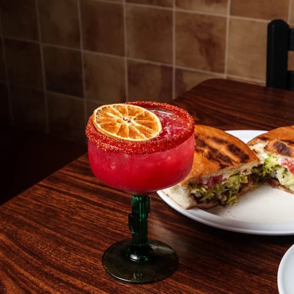 A vibrant red cocktail with a dried orange slice and chili-lime rim, next to a torta on a wooden table.